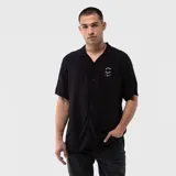 Camisa de manga corta color negro, con cuello solapa y cierre frontal de botones. Presenta un pequeño estampado circular en el pecho con texto en color blanco.