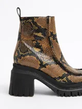 Botín de cuero con estampado de piel de serpiente en tonos ocre y negro, con puntera cuadrada, cierre lateral con cremallera, suela track de goma y tacón cuadrado.