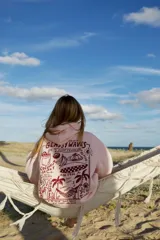 Hoodie rosa claro con estampado gráfico en la espalda en color rojo oscuro. El estampado incluye texto como "GLASSY WAVES SURF COMPANY", ilustraciones de bebidas, frutas, una tabla de surf y palmeras.