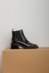 Botas tipo chelsea de cuero color marrón oscuro con acabado brillante. Presentan punta cuadrada con detalle de puntera metálica, elásticos laterales y taco cuadrado bajo.