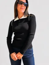 Chomba de tejido acanalado negro con cuello polo en contraste color blanco y cierre frontal con tres botones. Presenta un diseño ajustado al cuerpo con mangas largas y detalles de costuras blancas en los hombros.