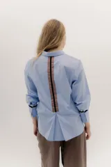 Camisa de manga larga color celeste claro, con cuello camisero y botones frontales. Presenta un detalle de cinta negra alrededor del puño doblado.