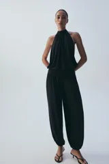 Mono largo negro con cuello halter, hombros descubiertos, parte superior drapeada y piernas anchas con bajo acabado en puño elástico.