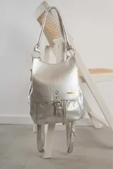 Mochila de cuero color plateado con diseño de solapa frontal, cierre de hebilla decorativa y correas ajustables. Cuenta con herrajes metálicos y una placa con logo en la parte frontal.