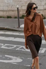 Camisa de lino color beige, con cuello solapa, escote en V y mangas largas. Presenta un diseño de bajo asimétrico con pliegues delanteros que generan un efecto abullonado.