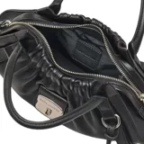 Cartera satchel compacta de cuero de cordero negro, con diseño estructurado, frunces en la parte superior, doble asa de mano y correa ajustable para llevar al hombro o cruzada. Incluye cierre metálico frontal con logo grabado.