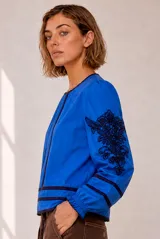 Blusa azul con cuello redondo, mangas largas abullonadas y puños elásticos fruncidos. Presenta bordados florales en negro en las mangas y detalles de ribetes oscuros en cuello, hombros y ruedo.