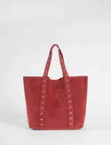 Cartera tipo shopper de gran tamaño confeccionada en material efecto gamuza color rojo. Presenta correas reforzadas decoradas con tachas metálicas plateadas y un dije colgante en forma de corazón con detalles de tachas. Cuenta con una placa metálica pequeña con logo en la parte inferior frontal.