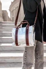 Cartera plateada tipo tote bag con detalles en cuero marrón, doble asa de mano y correa larga ajustable.