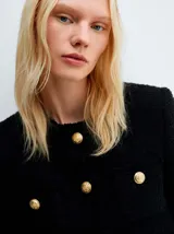 Chaqueta de estilo tweed en color negro, con cuello redondo, cierre frontal de botones dorados y bolsillos delanteros con solapa. Presenta un diseño de silueta recta y acabado deshilachado en los bordes.