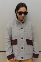 Campera de paño en tono gris melange, con cuello alto y cierre frontal de botones grandes. Presenta detalles en contraste de color marrón oscuro en los puños y en las solapas de los bolsillos frontales.