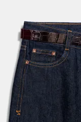 Jeans de tiro alto color azul oscuro, con cinco bolsillos, pernera ancha y nervio marcado en el delantero. Incluye un cinturón fino de cuero sintético marrón con textura de cocodrilo. Cierre frontal con cremallera y botón.