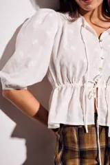 Blusa negra de algodón con diseño de broderie, mangas tres cuartos abullonadas, cierre frontal con botones y detalle de cordón ajustable en la cintura.