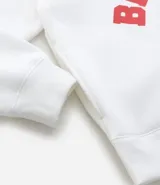 Hoodie blanco con capucha y cordones ajustables, confeccionado en algodón con felpa interior. Presenta estampado frontal con la palabra 'BROOKLYN' en color rojo, un parche circular con la imagen de Notorious B.I.G. y una firma bordada. Cuenta con bolsillo canguro y puños ajustados.