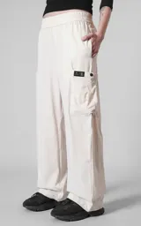 Pantalón cargo de corte oversize color beige, confeccionado en microfibra técnica. Presenta cintura elástica, bolsillos laterales y un bolsillo cargo con cierre de broche snap en el muslo. Incluye un sistema de elásticos con tancas en los tobillos para regular la apertura.