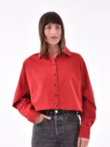 Camisa cropped color rojo con cuello clásico, botones negros en el frente y mangas largas con puños abotonados. Presenta un detalle de abertura en la parte superior de las mangas.