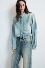Cazadora corta de denim en tono celeste claro, con cuello solapa, manga larga y cierre frontal con botones. Presenta un bolsillo de plastrón en el delantero y detalles de rotos en el cuello y el ruedo deshilachado.