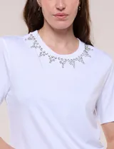 Remera blanca de calce recto y cuello redondo, confeccionada en suave jersey con detalle de aplicación de piedras brillantes a modo de gargantilla en el escote.