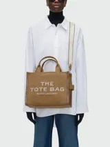 Bolso tipo tote de lona color negro con letras blancas bordadas en el frente que dicen 'THE TOTE BAG MARC JACOBS'. Cuenta con asas superiores cortas y una correa de hombro ajustable y extraíble.