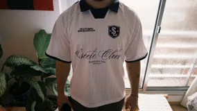 Camiseta de fútbol estilo boxy color blanco, con cuello polo y ribetes negros en las mangas. Presenta detalles estampados y bordados exclusivos en el frente y hombros.