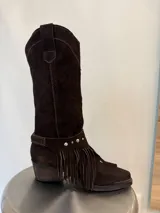 Bota de caña alta estilo western confeccionada en cuero vacuno color marrón oscuro. Presenta un diseño versátil con estribo desmontable decorado con flecos y tachas metálicas. Cuenta con taco de madera y punta fina.