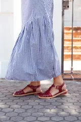 Sandalias planas estilo romano tipo cangrejera, confeccionadas en cuero charolado de color bordó. Presentan diseño de tiras entrelazadas en el empeine, puntera cuadrada, talón cerrado con tira ajustable mediante hebilla metálica dorada y suela plana con pequeño tacón.