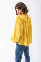 Camisa de corte holgado en color amarillo vibrante, confeccionada en tela fluida. Presenta cuello alto con volados, detalle de frunces en el canesú y mangas largas abullonadas con puños elásticos.