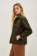 Campera estilo militar o safari color verde oliva, confeccionada en algodón. Presenta cuello solapa y bolsillos frontales con tapa. Destaca por sus apliques de pedrería o bordados en tonos marrones y dorados en el cuello y sobre los bolsillos.