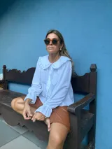 Camisa blanca de corte holgado con cuello bebé con volados, cierre frontal de botones y mangas largas con puños terminados en volados.