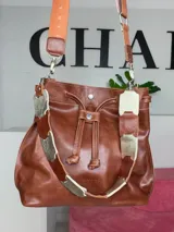 Bolso tipo bucket bag Chabas modelo 3.1, confeccionado en cuero genuino de color marrón. Cuenta con cierre de cordón ajustable, dos tipos de asas (una corta de cuero con detalles decorativos y otra larga tipo bandolera con tira textil) y diseño sin forro interior.