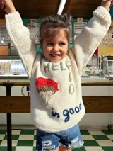 Sweater tejido a mano en lana merino color crema, con cuello redondo y mangas largas. Presenta un aplique bordado de un helado de dos bochas y texto bordado con la frase "Help is good".