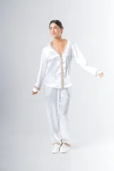 Pijama blanco de satén elastizado, compuesto por una camisa de manga larga con escote en V, abotonada adelante y con puntilla en escote y puños, y un pantalón recto con bolsillos laterales, elástico en la cintura y puntilla en el ruedo.