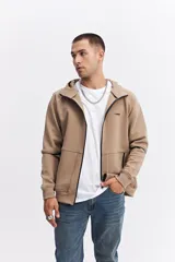 Campera con capucha y cierre frontal, color beige, con detalles de costuras y cierres en negro. Presenta mangas largas con puños acanalados y logo pequeño en el pecho.