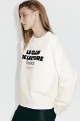 Buzo oversize de color beige con cuello redondo, hombros caídos y puños y bajo acanalados. Cuenta con un estampado frontal con texto en contraste.