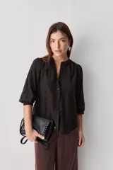 Camisa negra de textura liviana con detalle de lunares en relieve, cuello mao y cierre frontal con botones. Presenta mangas 3/4 con puños elastizados y frunces en los hombros.