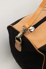 Bolso de viaje tipo duffel de lona negra con detalles en cuero tostado, asas de mano y correa ajustable para el hombro.