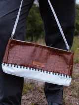 Sobre tipo baguette de cuero con textura croco en color marrón. Presenta un diseño bicolor con un lateral en cuero blanco unido mediante un trenzado artesanal en color negro. Incluye una etiqueta de cuero con el logo de la marca y un lazo desmontable.