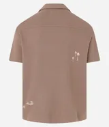 Camisa de manga corta, color marrón topo, con cuello camisero y corte amplio (boxy). Presenta bordados minimalistas en blanco en el frente, simulando un paisaje.
