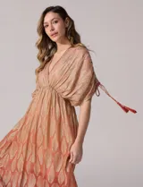 Vestido largo de gasa color salmón con un sutil patrón estampado brillante en tono dorado. Cuenta con escote en V cruzado, cintura imperio elastizada y mangas cortas con tiras ajustables terminadas en borlas.