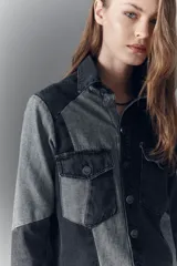 Campera de denim rígido con diseño de recortes en tonos gris y negro. Presenta cuello camisero, cierre frontal con botones metálicos, bolsillos con solapa en el pecho y corte recto.