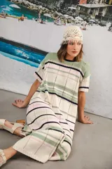 Vestido largo de tejido calado con rayas horizontales en tonos beige, marrón y amarillo. Tiene corte recto, hombros caídos, tajo lateral y logo bordado en la manga.