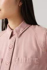 Camisa color rosa pálido, de corte cropped, con cuello camisero, cierre frontal con botones, mangas largas abullonadas con puños elásticos, dos bolsillos de parche en el pecho y canesú en la espalda.