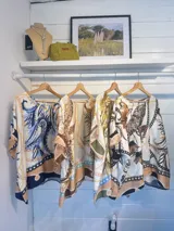 Blusa de satén con estampado de cadenas y hojas en tonos blanco, marrón y negro. Tiene corte amplio, mangas cortas y cuello redondo. Se ajusta a la cintura con un cinturón elástico negro con hebilla dorada.