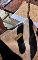 Cartera bandolera compacta de cuero con estampado animal print tipo leopardo, solapa frontal, costuras artesanales a la vista en los bordes y flecos decorativos colgantes. Cuenta con una manija larga trenzada y detalles de herrajes metálicos dorados.