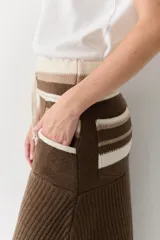 Falda midi de lana en color marrón, con diseño acampanado y tejido acanalado. Presenta una cintura elástica con cordón de ajuste, detalles de rayas en tonos beige y blanco en la parte superior, y costuras decorativas en el ruedo.