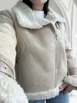 Chaqueta color beige con interior de piel sintética blanca, cierre cruzado con cremallera y detalles de hebillas en los puños.