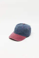 Gorra tipo baseball con diseño bicolor, cuerpo en azul desgastado y visera en color bordó. Presenta ojales de ventilación bordados en contraste y botón superior a tono con la visera.