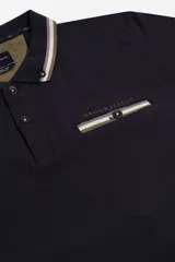 Remera polo azul marino de manga corta, con cuello y puños a rayas blancas y verde militar. Presenta un bolsillo en el pecho con detalle de franja a rayas y el logo de la marca "Brooksfield" bordado en dorado.