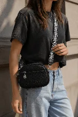Cartera bandolera negra de textura granulada, con bolsillo frontal con cierre y herrajes metálicos plateados. Incluye correa ajustable de tela con diseño bordado en blanco y negro.