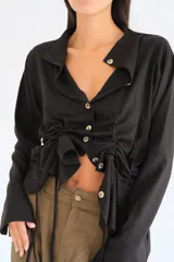 Camisa negra de lino con cuello camisero, manga larga y cierre frontal con botones de nácar. Presenta un diseño con cordones laterales que permiten fruncir la prenda para un ajuste personalizado.